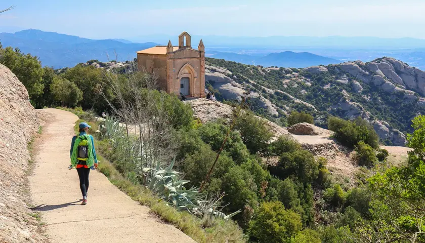 Une randonnée à Montserrat, avec une femme portant un sac à dos et une corde, menant à une petite chapelle perchée sur une colline rocheuse.