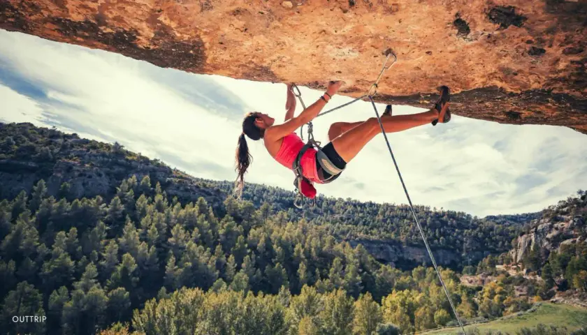 Une femme en pleine escalade extrême, suspendue sous un surplomb rocheux, avec une forêt verdoyante en arrière-plan.
