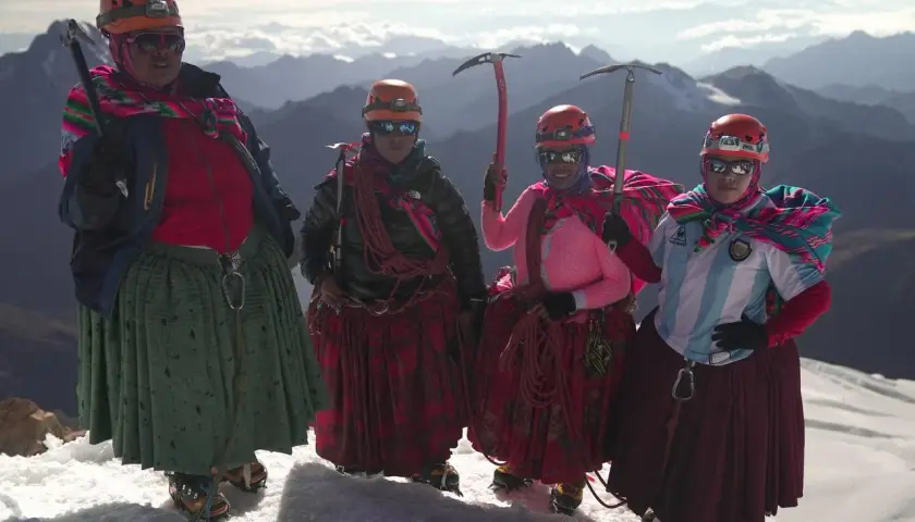 Quatre cholitas escaladoras, casques et piolets, posent fièrement sur un sommet enneigé, leurs jupes colorées flottant au vent.