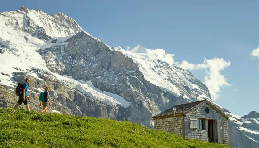 Deux randonneurs traversent une prairie verdoyante, avec un chalet en bois et les majestueux sommets de l'Eiger et de la Jungfrau en toile de fond.