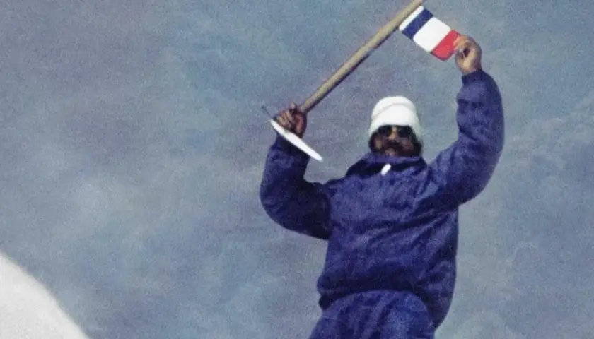Les mains de Maurice Herzog brandissent un drapeau français au sommet d'une montagne enneigée, symbole d'une victoire alpine.