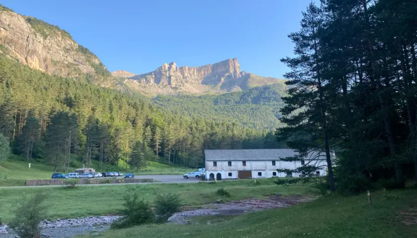 Vue panoramique de la **selva de Oza** avec une bâtisse blanche, des montagnes imposantes et une forêt dense sous un ciel bleu clair.