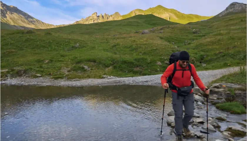 Un randonneur traverse un ruisseau en utilisant des bâtons de marche. Le soleil éclaire les montagnes verdoyantes en arrière-plan.