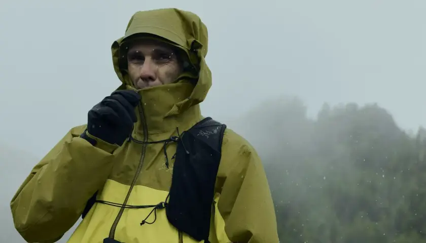 Un homme en veste Gore-Tex jaune et verte, sous la pluie, ajuste son col. Le lavage de cette veste est essentiel pour maintenir son imperméabilité.
