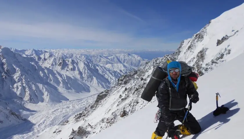 Daniele Nardi, alpiniste, en pleine ascension sur une pente enneigée, avec un sac à dos imposant et des crampons. Le paysage montagneux est spectaculaire.