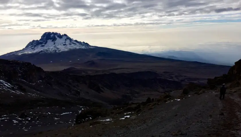 Vue sommet Kilimandjaro, paysage désertique et enneigé sous un ciel nuageux. Un randonneur solitaire sur le sentier.
