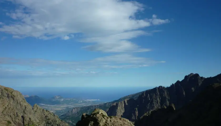 Vue spectaculaire sur la mer depuis les montagnes escarpées du GR20 partie nord. Le ciel bleu est parsemé de nuages blancs.