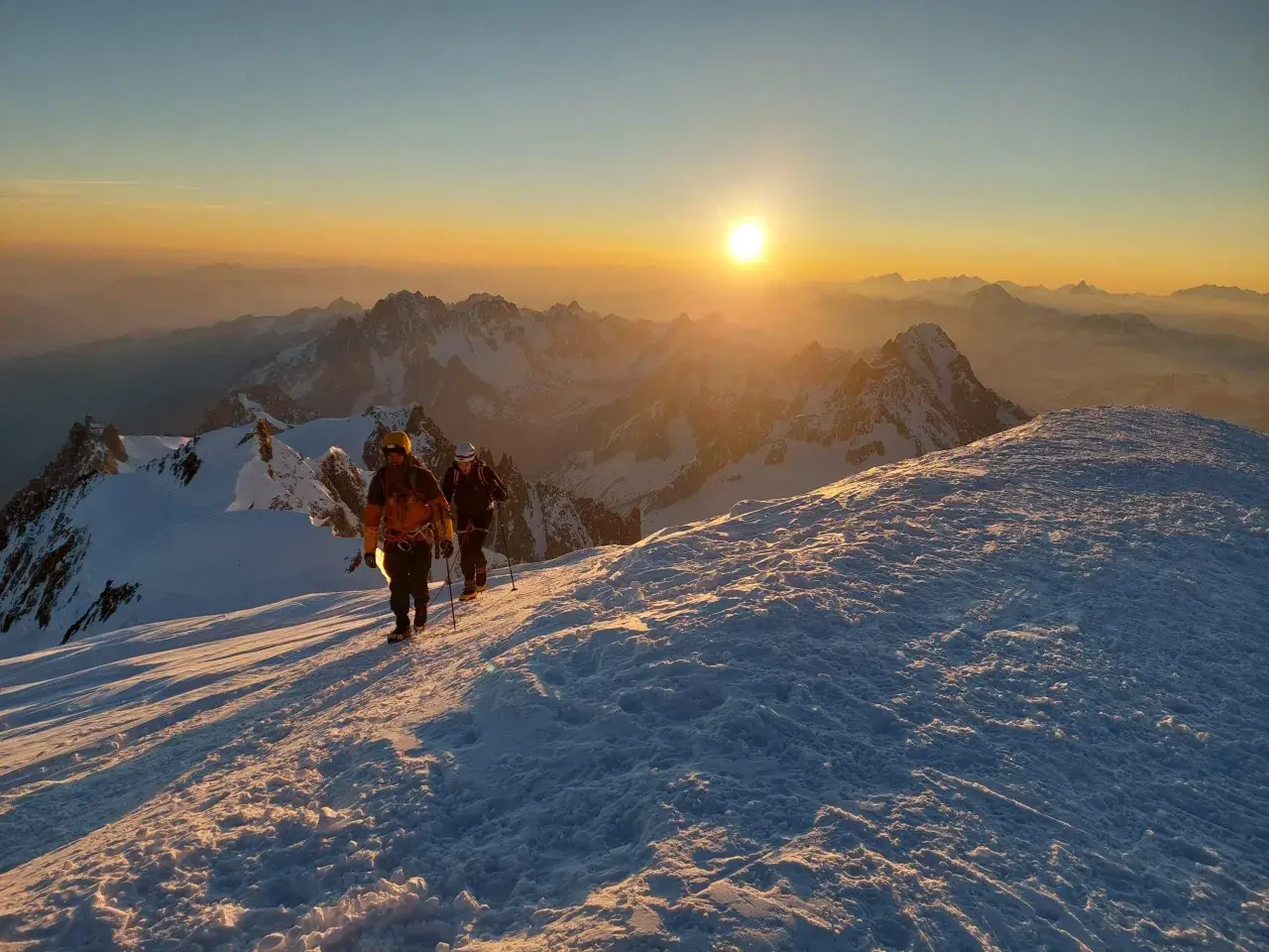 Deux alpinistes progressent sur un sentier enneigé au lever du soleil, dominant un paysage de montagnes spectaculaire. Le sommet montagneux est baigné d'une lumière dorée.