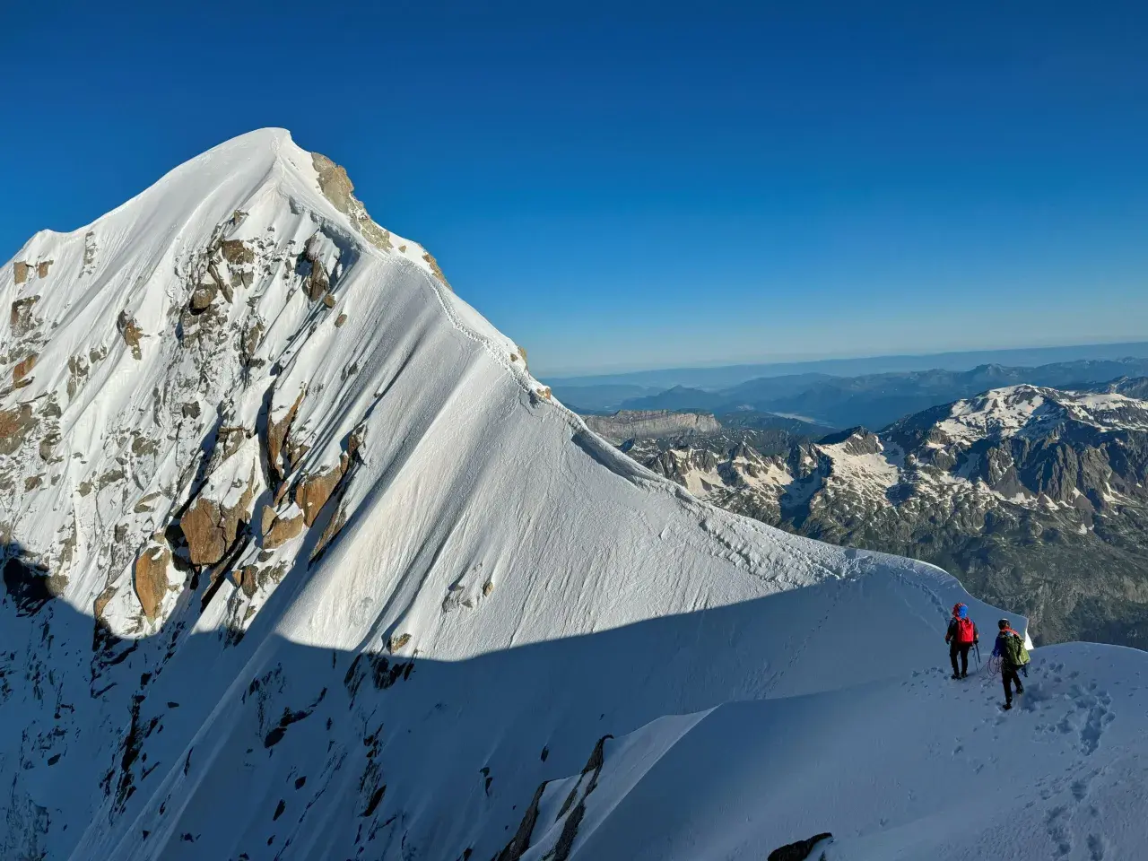 Deux alpinistes escaladent la crête enneigée de l'Aiguille Verte, sous un ciel bleu intense. Le soleil projette de longues ombres sur la montagne.