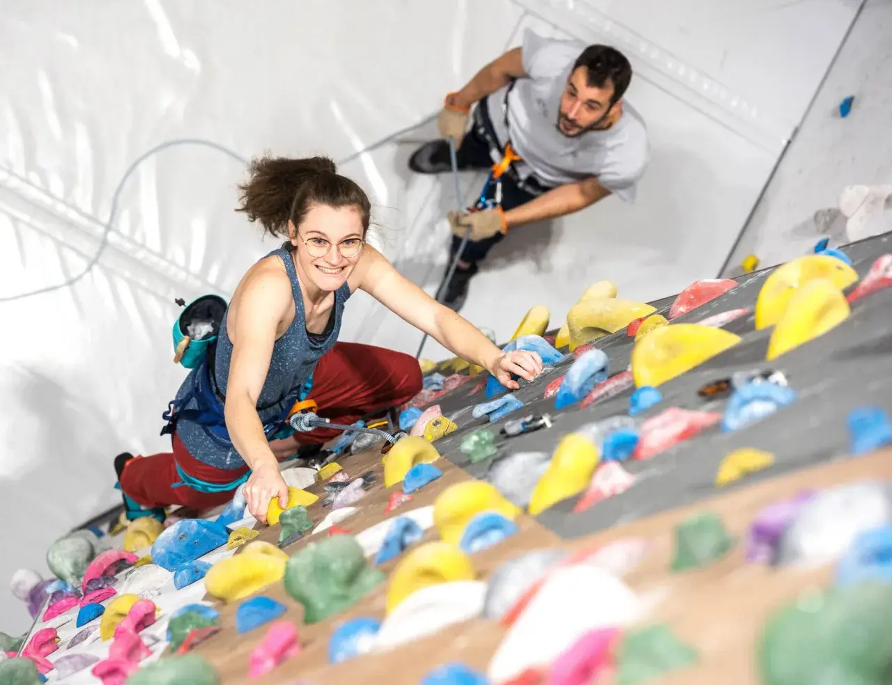 Une femme souriante en tenue d'escalade grimpe un mur coloré, assurée par un homme en arrière-plan.