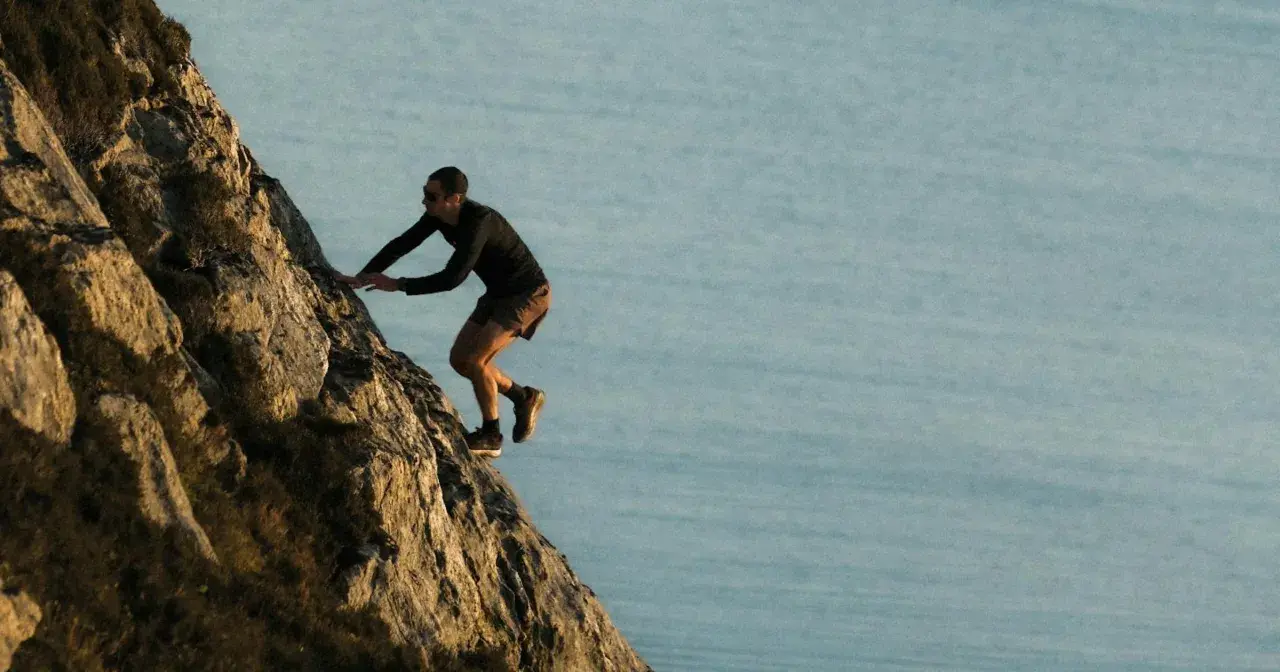 Kilian Jornet escalade une paroi rocheuse abrupte, dominant un plan d'eau calme, rappelant l'esprit d'aventure dans les Pyrénées.