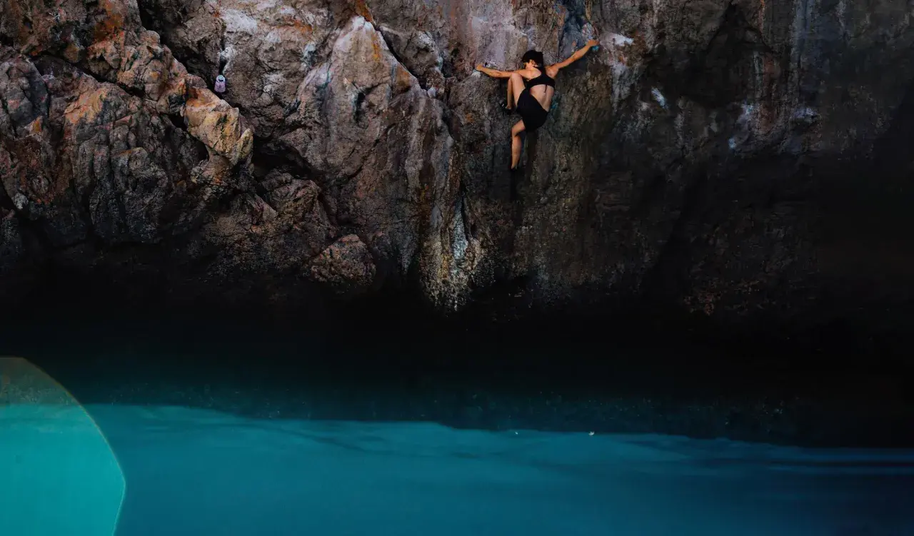 Une femme pratique l'escalade outdoor au-dessus d'une eau turquoise. Elle s'agrippe à la paroi rocheuse, concentrée sur sa prochaine prise.