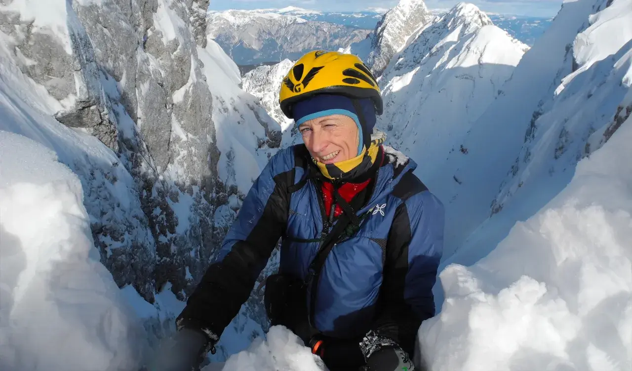 Une alpiniste souriante, Nives Meroi, en plein effort sur une crête enneigée, casque jaune, montagnes majestueuses en arrière-plan.