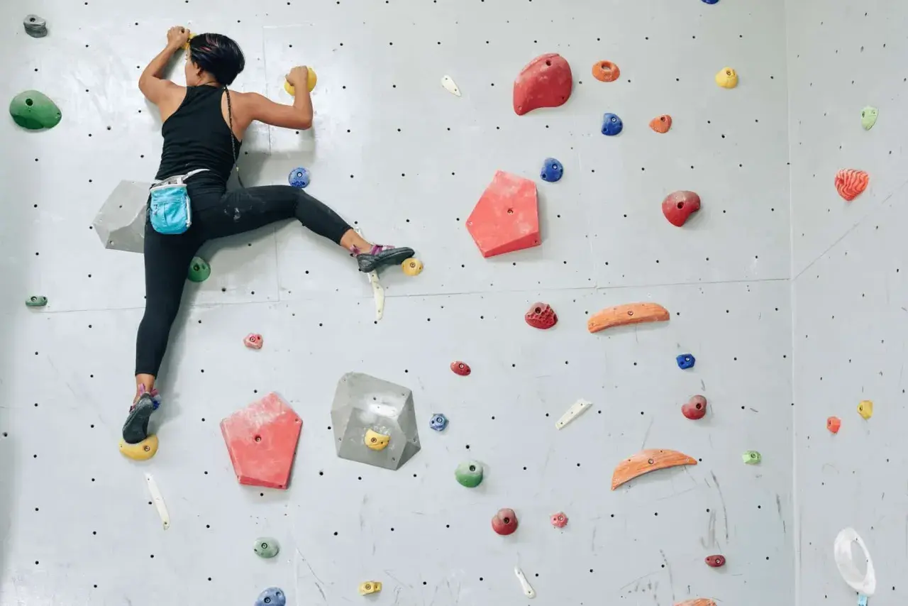 Grimpeuse en action, un pied dans la main, sur un mur d'escalade coloré. Elle progresse avec agilité.