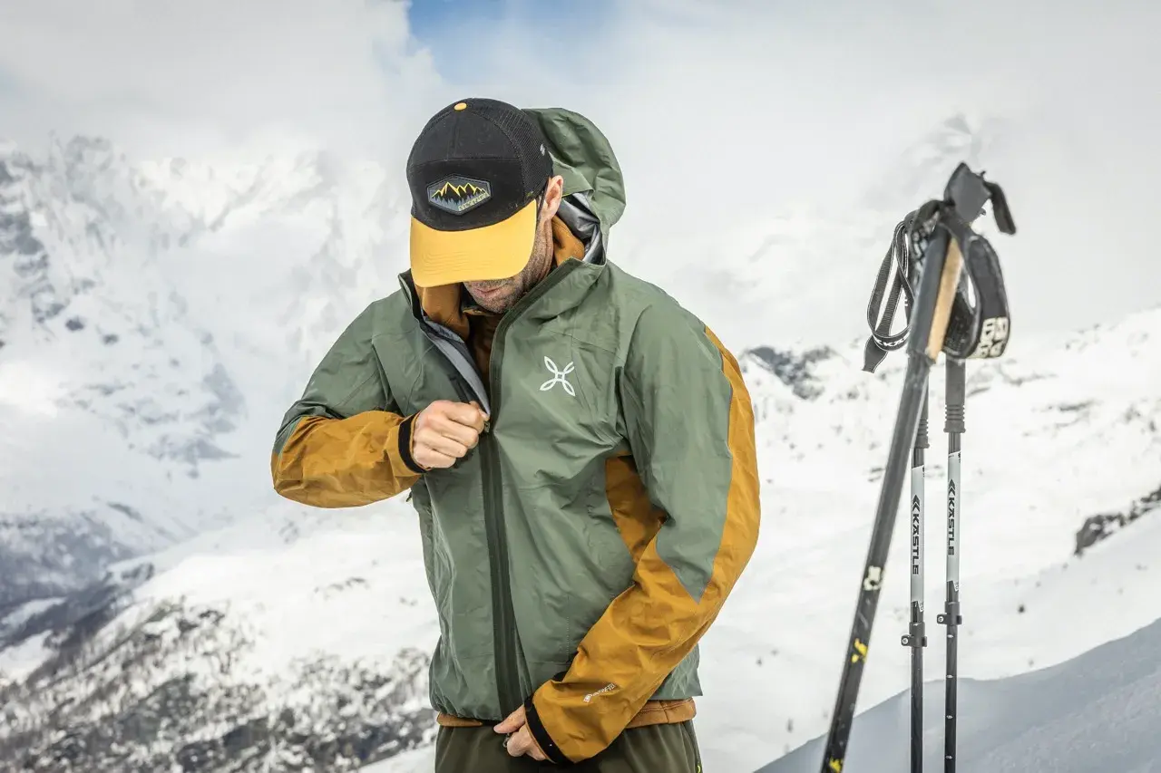 Un skieur ajuste sa veste technique, peut-être pour comparer le confort entre Primaloft vs Gore-Tex, avec des montagnes enneigées en arrière-plan.