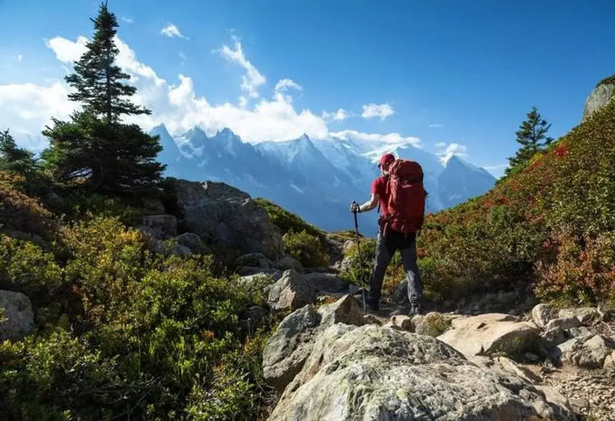Un randonneur solo, sac à dos rouge, bâtons de marche, admire les montagnes majestueuses sous un ciel bleu éclatant.