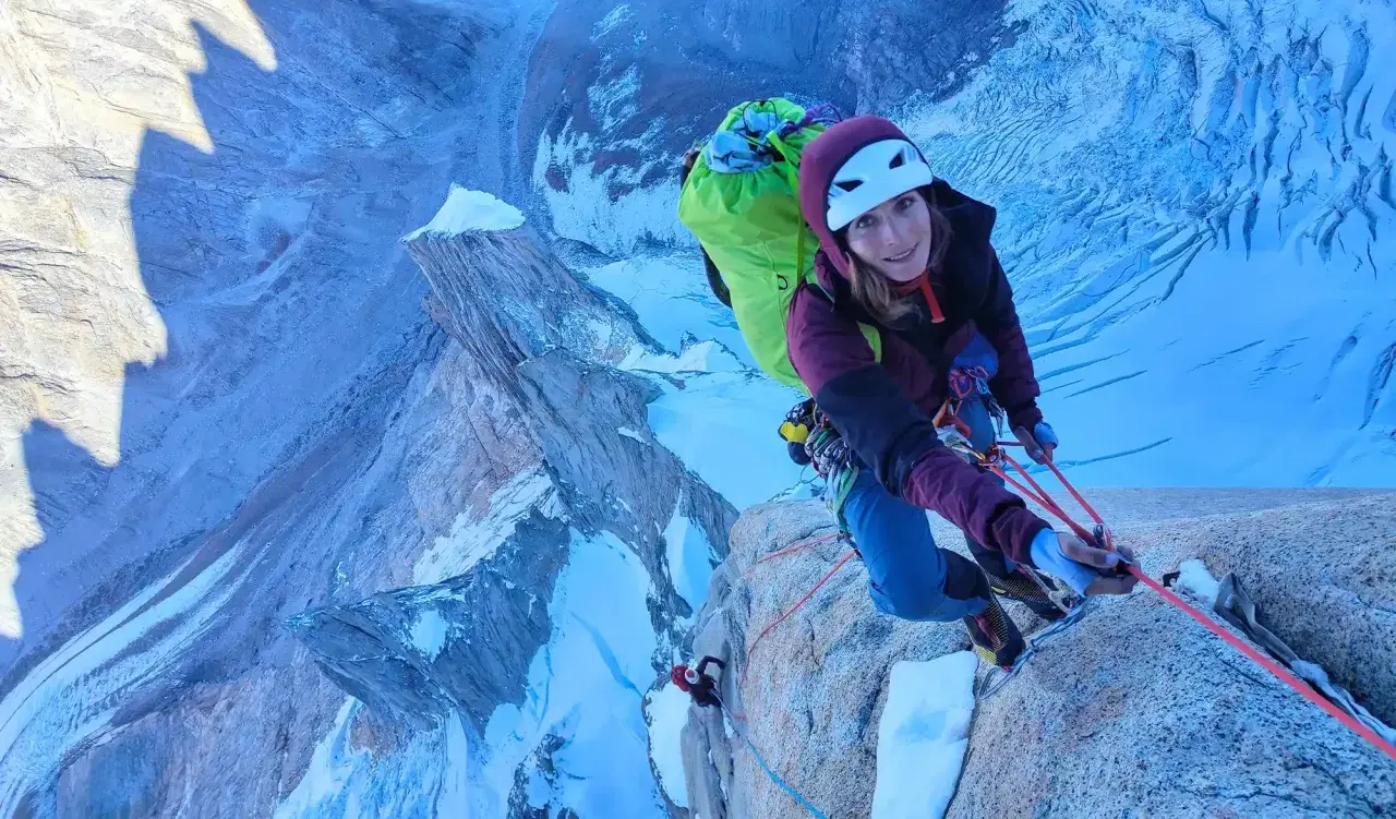 Lise Billon, casque blanc, grimpeuse souriante, est en pleine ascension sur une paroi rocheuse glacée, avec un sac à dos vert vif.