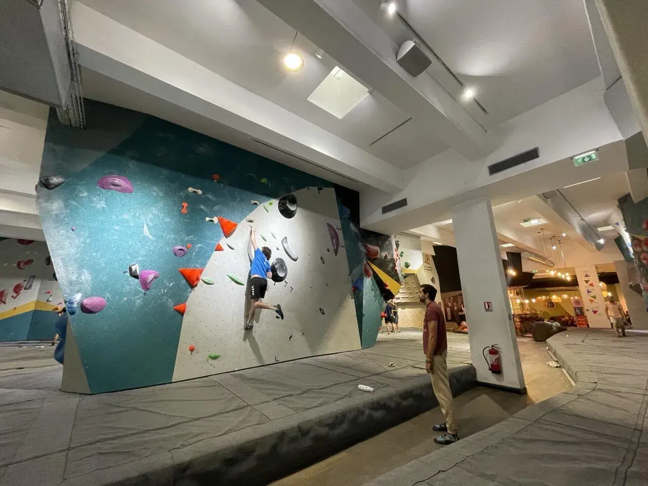 Un jeune homme en bleu pratique l'escalade sur un mur coloré, un autre spectateur l'observe. L'ambiance est propice à l'escalade nation.