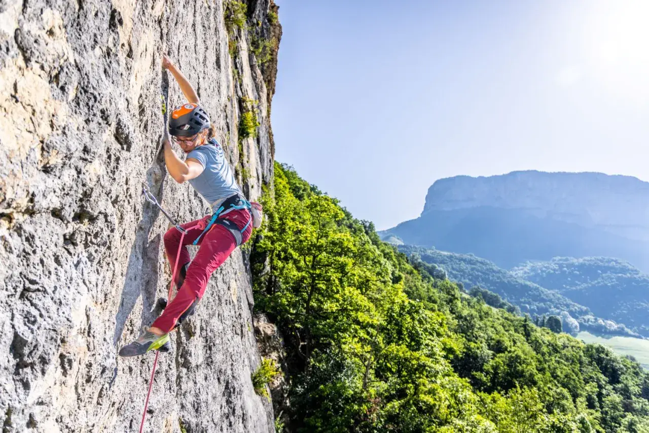 Une grimpeuse évolue sur une paroi rocheuse, profitant d'une journée ensoleillée dans ce parc d'escalade naturel. Le paysage montagneux verdoyant offre un panorama spectaculaire.