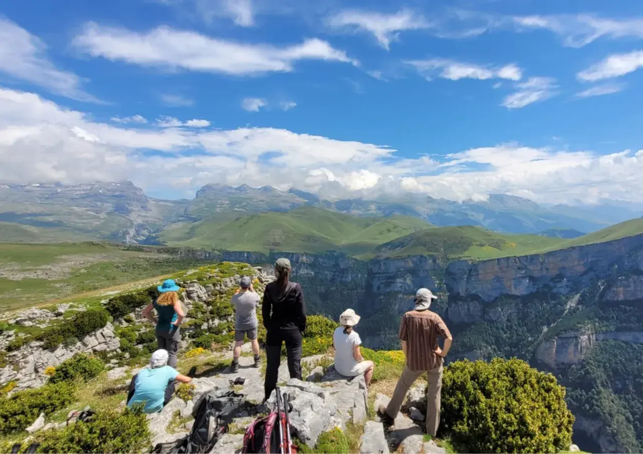 Randonneurs contemplant le paysage spectaculaire du **parque nacional de Ordesa y Monte Perdido**, avec ses falaises abruptes et ses montagnes verdoyantes sous un ciel bleu.