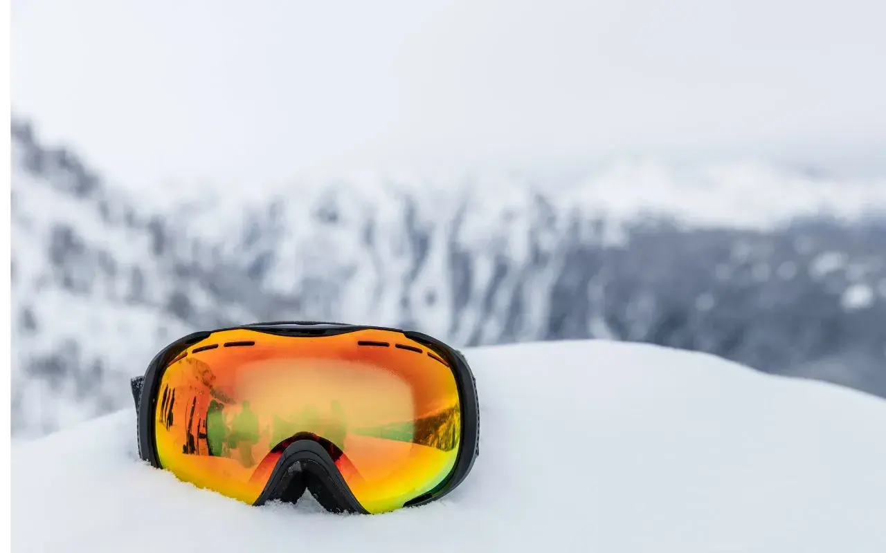 Masque de ski orange posé dans la neige, reflétant des skieurs. Indispensable pour savoir quel masque de ski choisir.