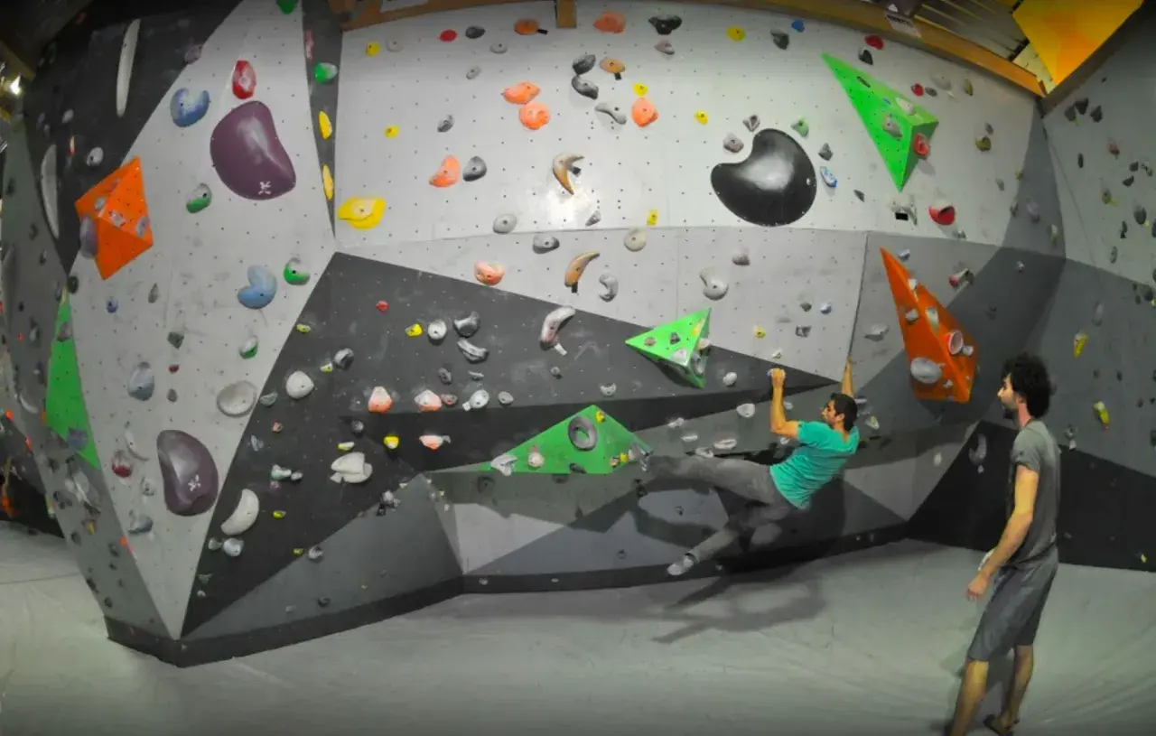 Un homme en t-shirt vert fait de l'escalade pertuis sur un mur coloré, un autre homme le regarde.