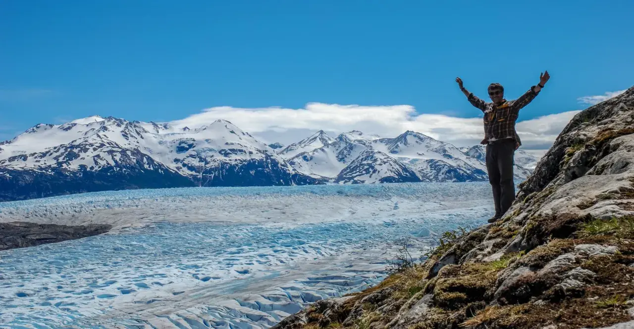 Un homme bras levés devant un glacier immense et des montagnes enneigées de la Patagonie.