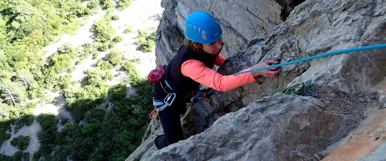 Une grimpeuse en casque bleu progresse sur une paroi rocheuse, corde bleue en main. L'escalade à Thaurac offre des vues spectaculaires.