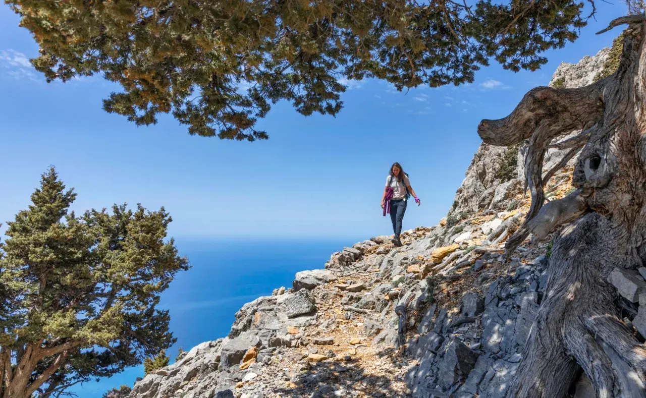 Une randonneuse explore un sentier rocailleux sur une montagne de Crète, avec la mer bleue en arrière-plan.