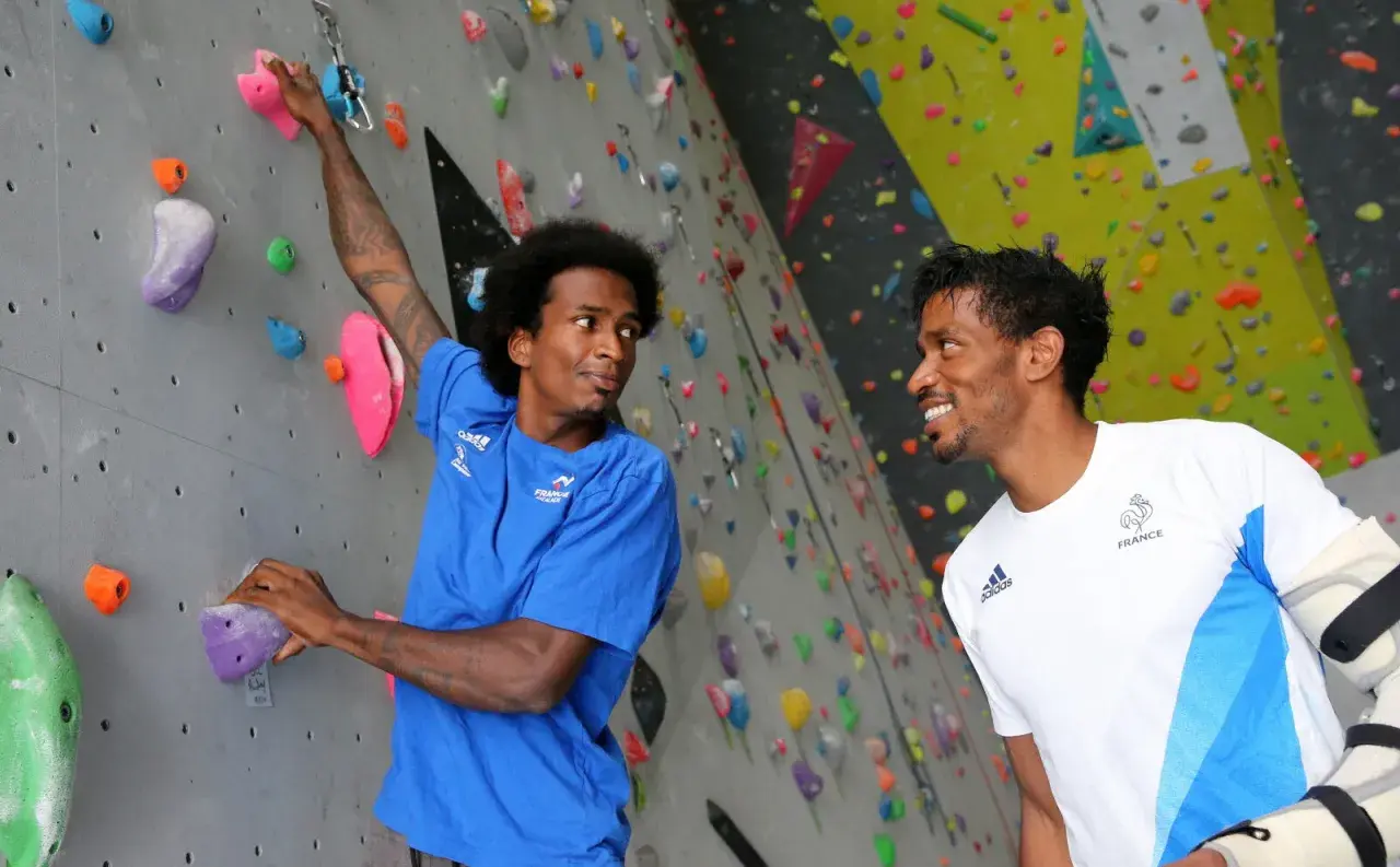 Deux grimpeurs, dont frere mawem, sourient sur un mur d'escalade coloré. L'un est en bleu, l'autre en blanc avec le logo de la France.