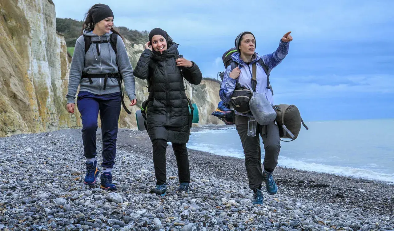 Trois randonneuses marchent sur une plage de galets, le ciel est couvert. L'une d'elles pointe le doigt vers l'horizon, peut-être pour indiquer la fin de leur GR 21 étapes.