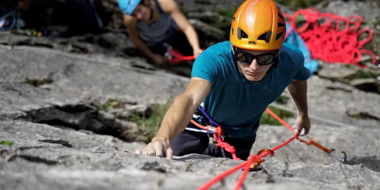 Un grimpeur concentré, casque orange, s'engage dans une voie d'escalade 64. Une corde rouge vif l'assure.