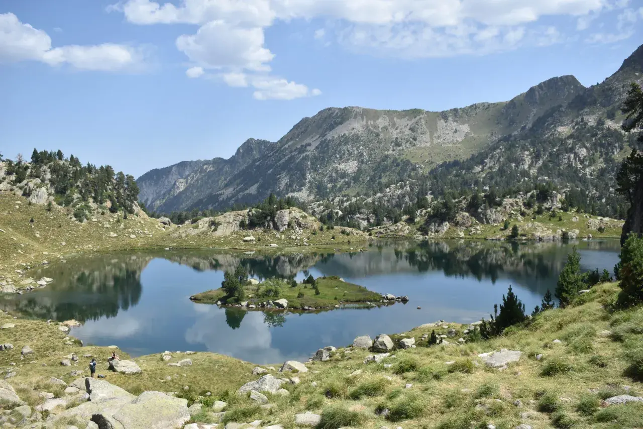 Vue paisible du cirque de Colomers, avec un lac miroir reflétant les montagnes et une petite île verdoyante.