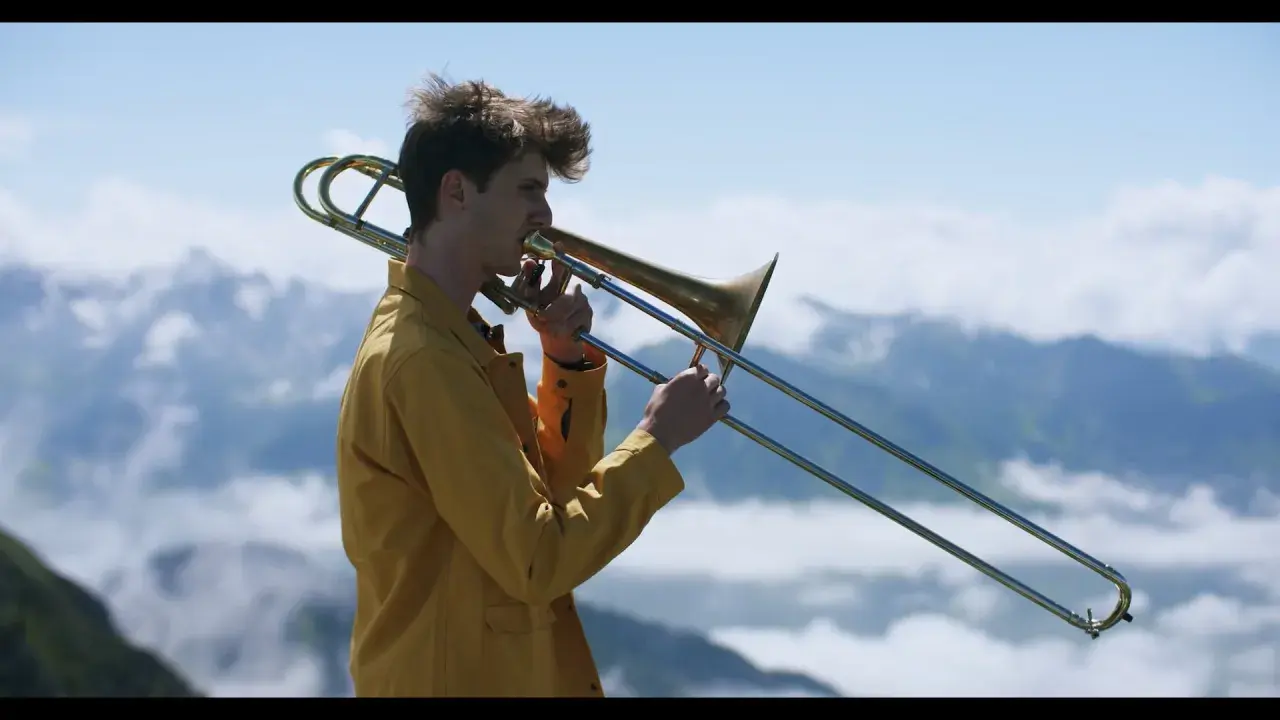 Victor Decamp Destivelle, un jeune homme aux cheveux ébouriffés, joue du trombone dans un paysage montagneux brumeux.