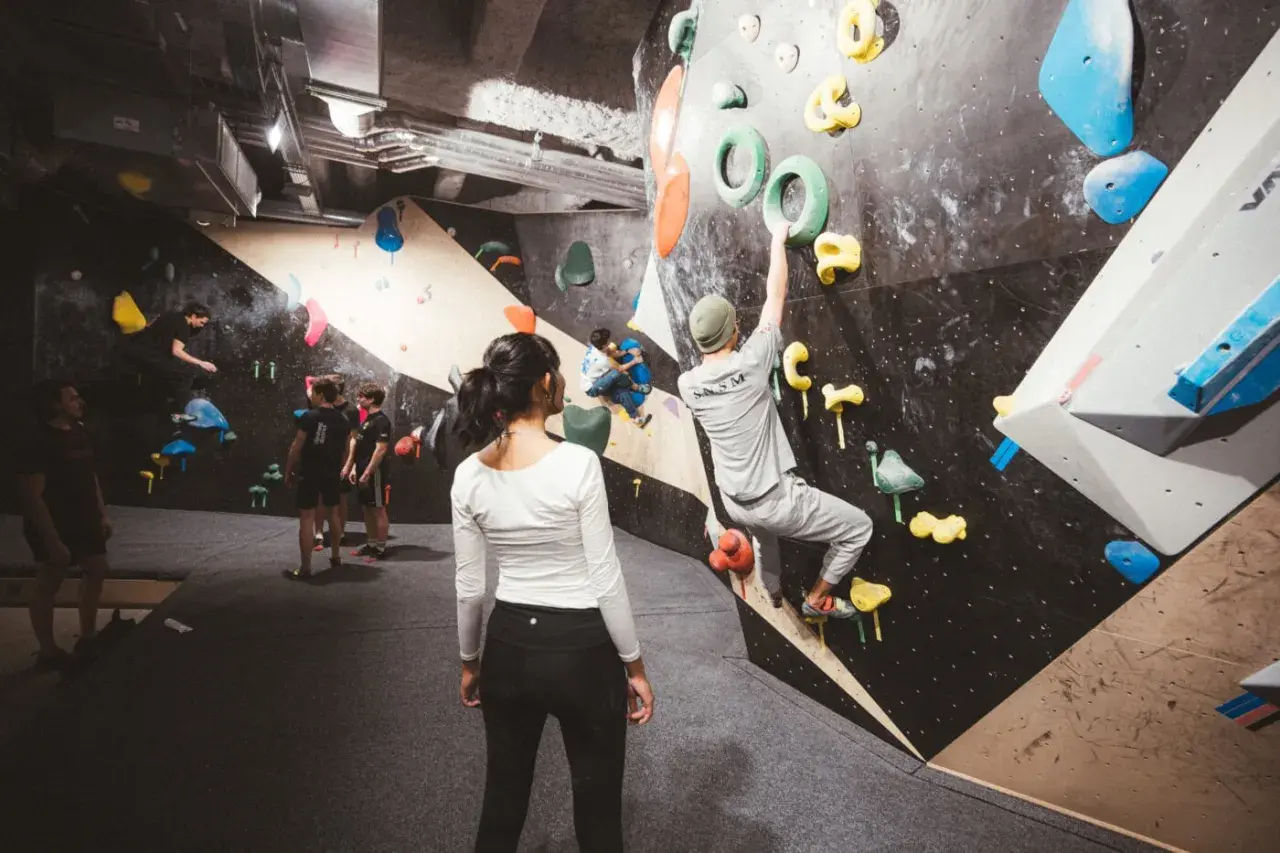 Des grimpeurs s'entraînent dans une salle de block animée, certains escaladent des murs colorés, d'autres observent.