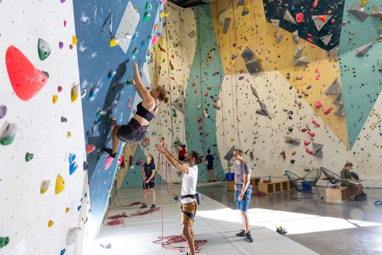 Une femme fait de l'escalade climb up sur un mur coloré, d'autres personnes observent ou se préparent.