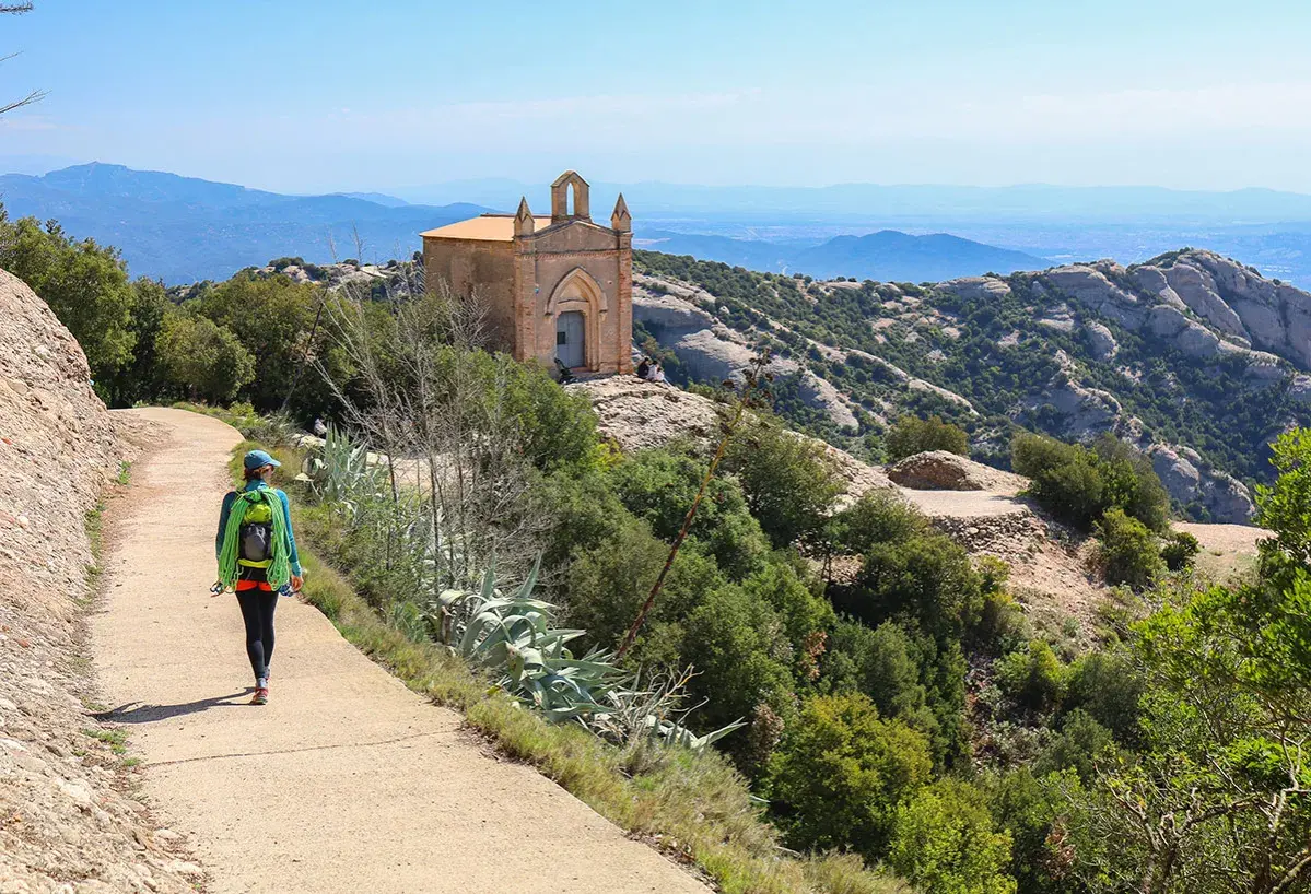Une randonnée à Montserrat, avec une femme portant un sac à dos et une corde, menant à une petite chapelle perchée sur une colline rocheuse.