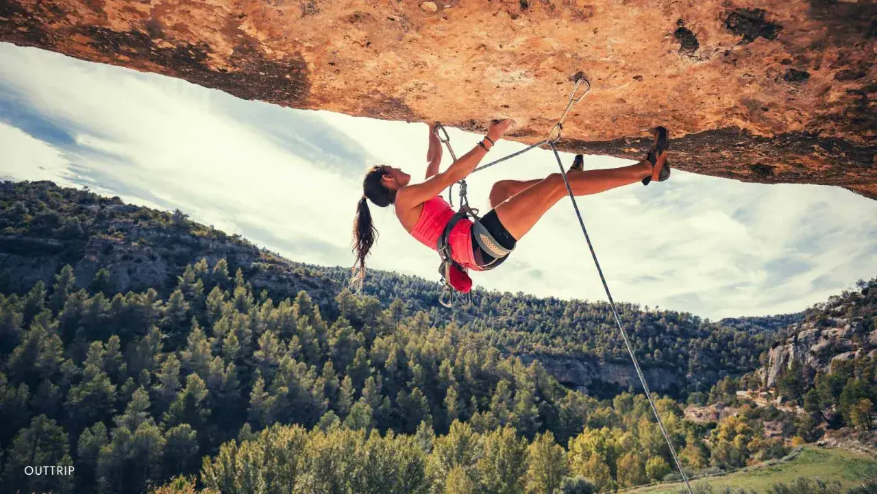 Une femme en pleine escalade extrême, suspendue sous un surplomb rocheux, avec une forêt verdoyante en arrière-plan.