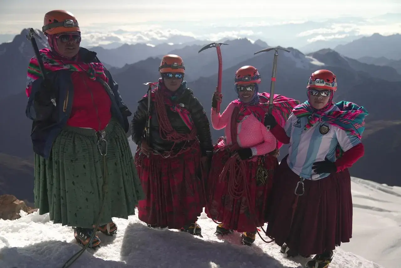 Quatre cholitas escaladoras, casques et piolets, posent fièrement sur un sommet enneigé, leurs jupes colorées flottant au vent.