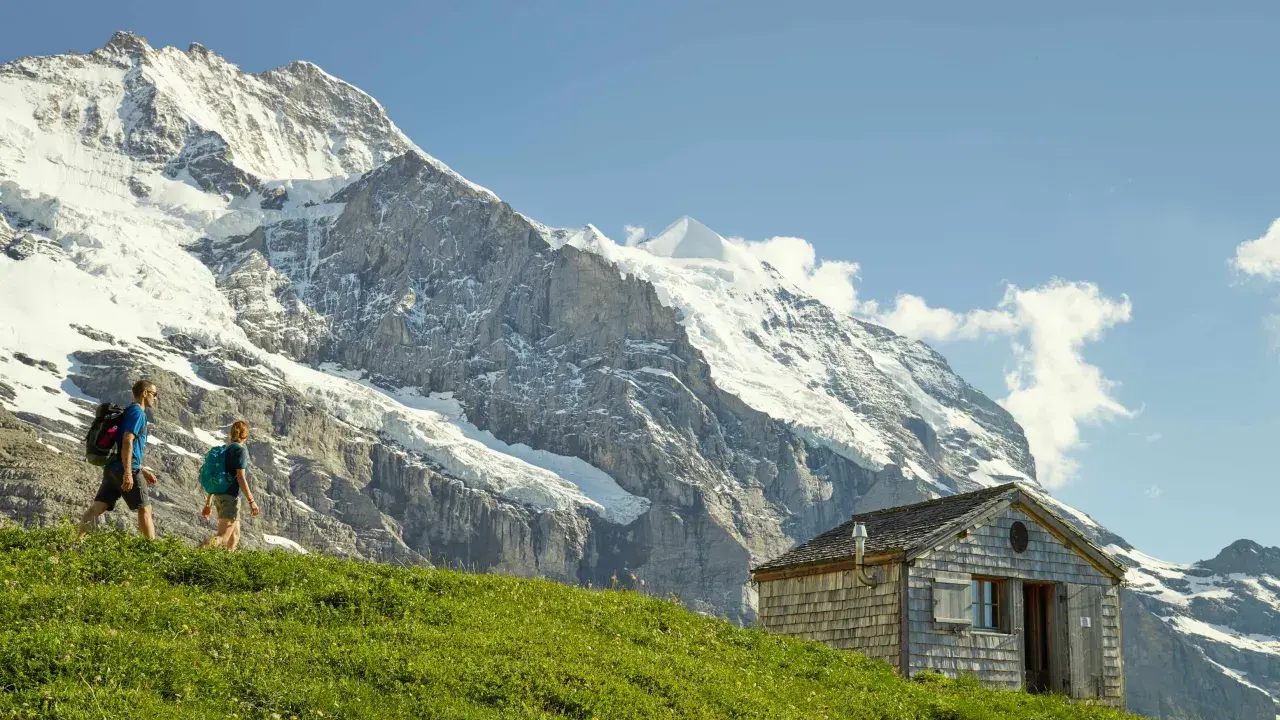 Deux randonneurs traversent une prairie verdoyante, avec un chalet en bois et les majestueux sommets de l'Eiger et de la Jungfrau en toile de fond.