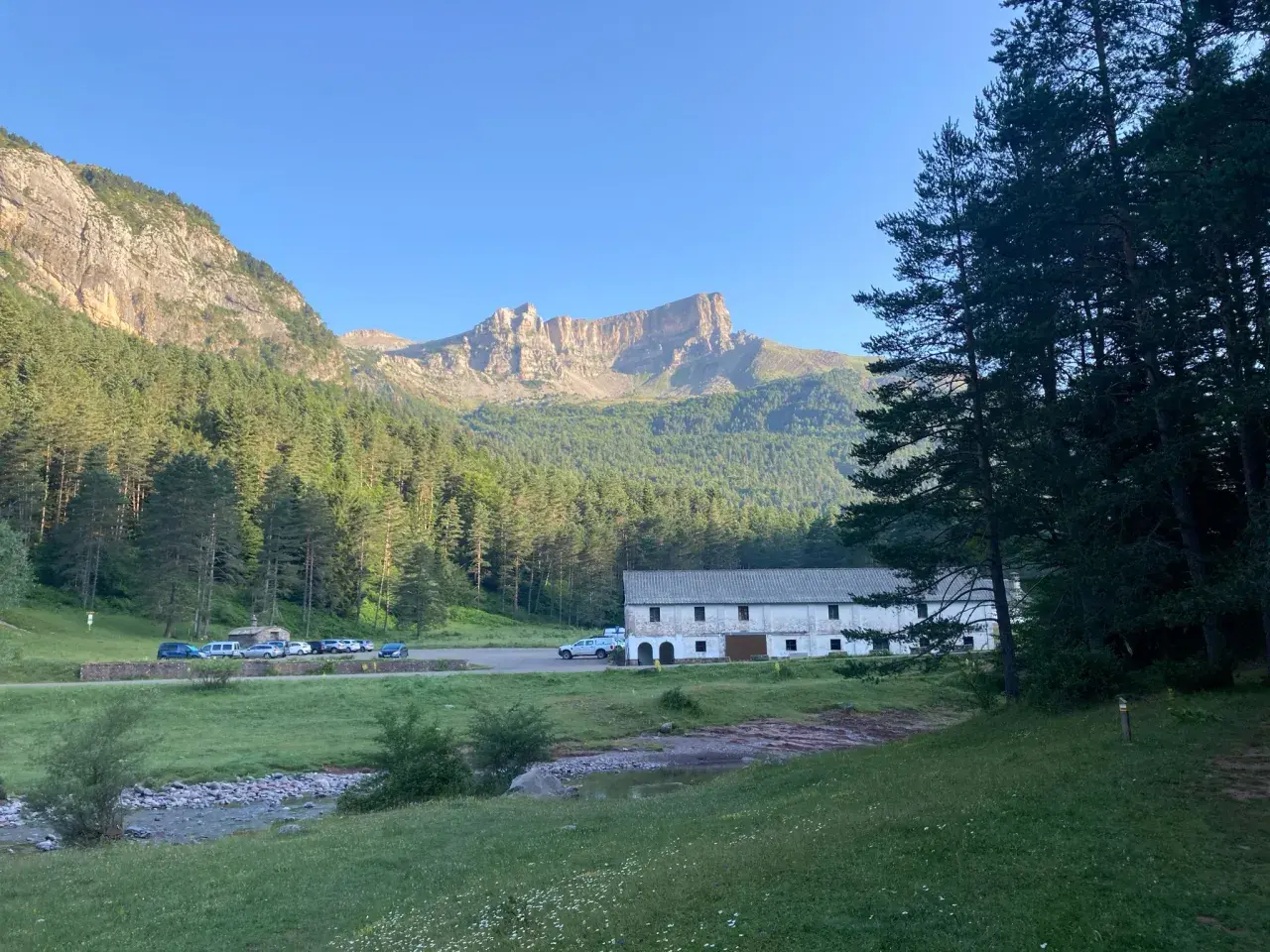 Vue panoramique de la **selva de Oza** avec une bâtisse blanche, des montagnes imposantes et une forêt dense sous un ciel bleu clair.