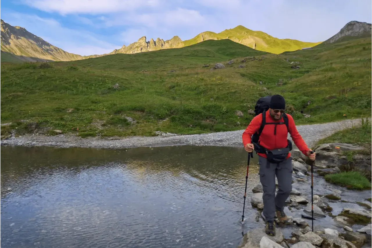 Un randonneur traverse un ruisseau en utilisant des bâtons de marche. Le soleil éclaire les montagnes verdoyantes en arrière-plan.