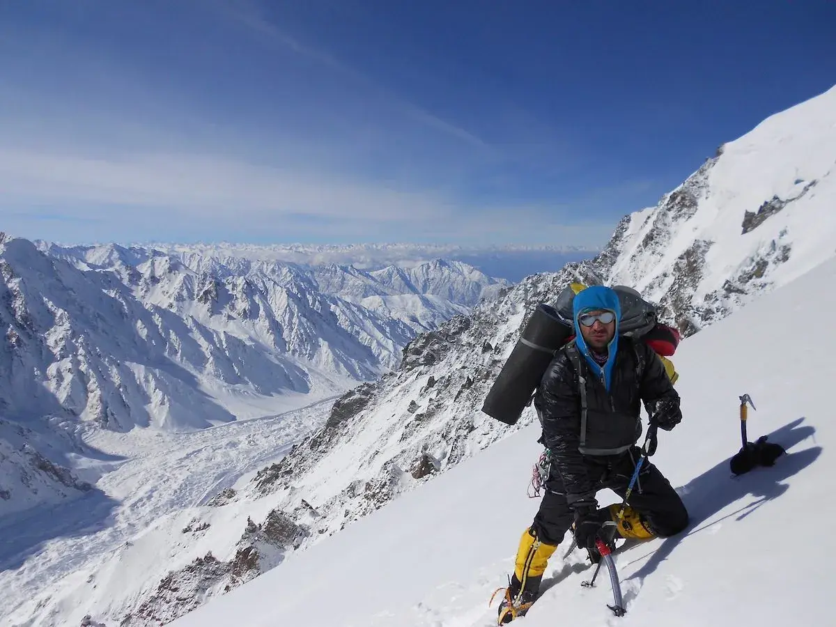Daniele Nardi, alpiniste, en pleine ascension sur une pente enneigée, avec un sac à dos imposant et des crampons. Le paysage montagneux est spectaculaire.