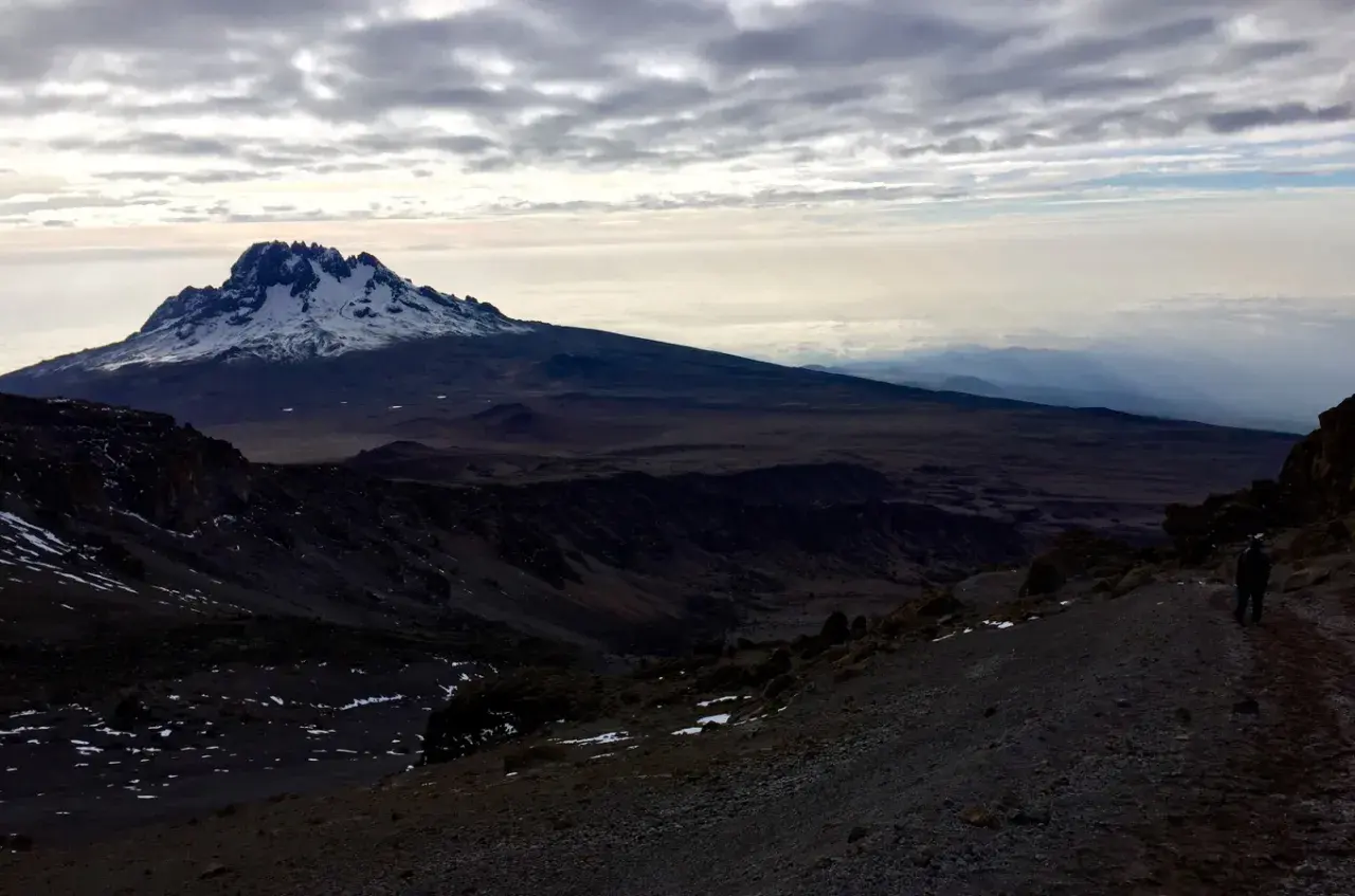 Vue sommet Kilimandjaro, paysage désertique et enneigé sous un ciel nuageux. Un randonneur solitaire sur le sentier.