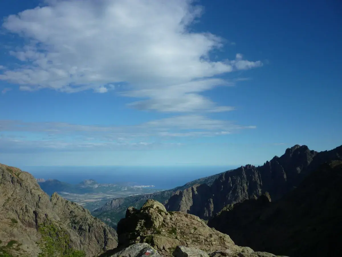 Vue spectaculaire sur la mer depuis les montagnes escarpées du GR20 partie nord. Le ciel bleu est parsemé de nuages blancs.