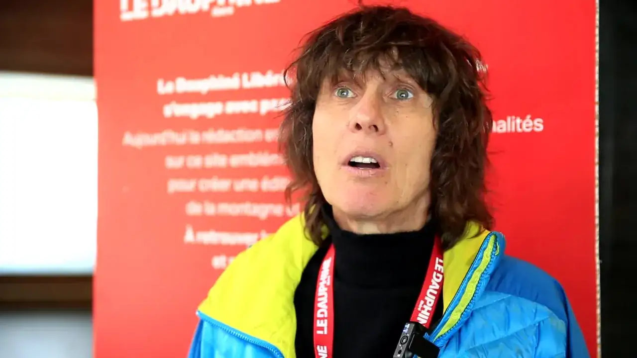 Catherine Destivelle aujourd'hui, regard intense, porte une veste bleue et jaune. Elle est devant un panneau rouge avec du texte.