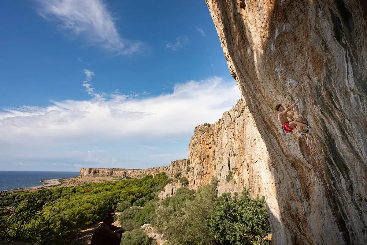 Un grimpeur d&eacute;fie une paroi rocheuse spectaculaire en Sicile, avec la mer et une v&eacute;g&eacute;tation luxuriante en arri&egrave;re-plan.