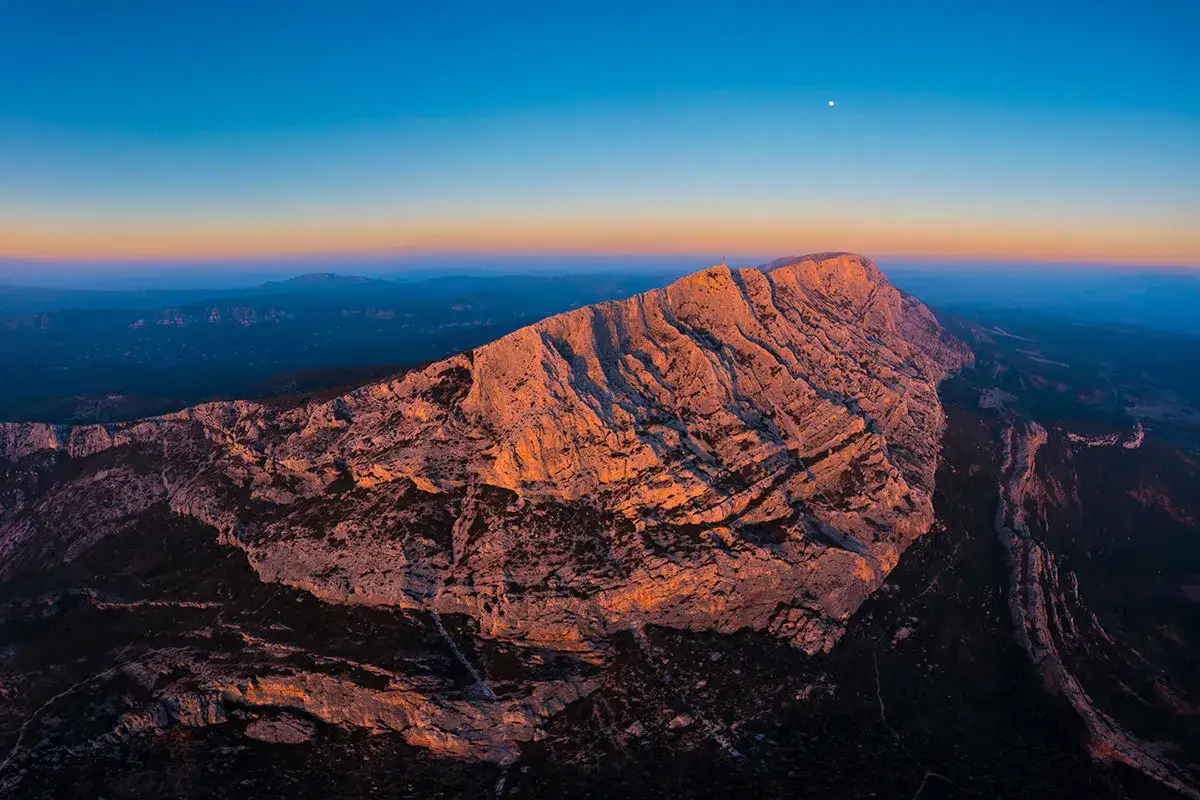 Le soleil couchant illumine la montagne Sainte-Victoire, offrant un spectacle grandiose pour l'escalade. La lune veille sur ce paysage.