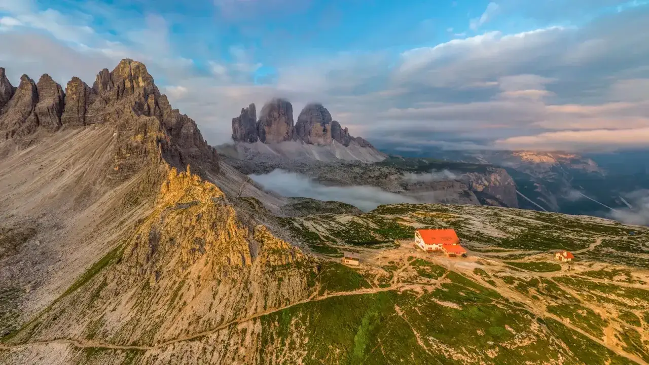 Refuge sur l'Alta Via 2 des Dolomites, avec les Tre Cime di Lavaredo &eacute;mergeant des nuages. Un sentier serpente sur les pentes verdoyantes.