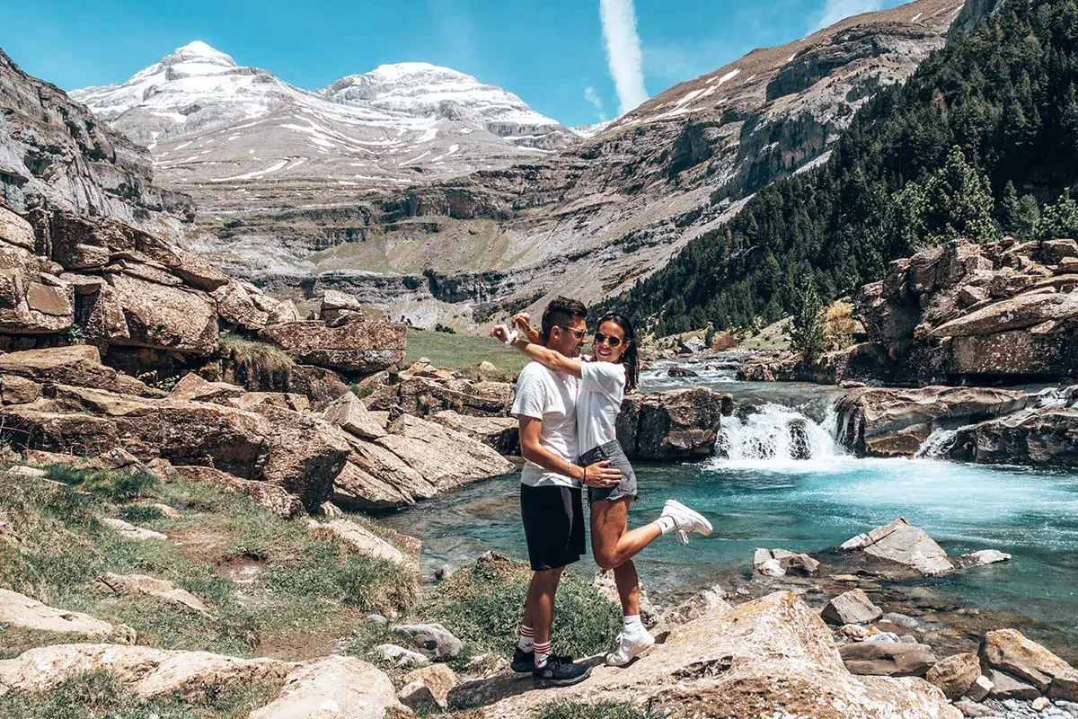 Couple s'embrassant près d'une rivière aux eaux turquoise dans le parc national d'Ordesa y Monte Perdido, avec des montagnes enneigées en arrière-plan.